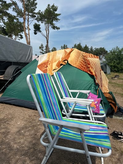 In the foreground of the image, two colorful striped folding chairs sit on a dirt and gravel campsite. Behind the chairs is a green tent with a patterned blanket draped over its top. To the left of the tent, a portion of a gray tarp-covered trailer is visible. Trees and a blue sky with scattered clouds form the background. The scene depicts an outdoor campsite, likely in the city of Kolodezny, Russia.