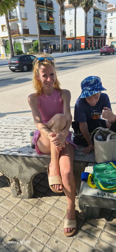 In Torre Del Mar, Spain, a woman with blonde hair pulled back and sunglasses perched on her head sits on a concrete bench, leaning forward with her legs crossed. She is wearing a pink sleeveless top and a pink skirt. To her right, a young boy wearing a blue tie-dye bucket hat and a blue t-shirt sits looking down. They are positioned on a tiled walkway in front of a building with palm trees. In the background, buildings with balconies and awnings are visible, along with parked cars on a street. A green and yellow soccer-themed bag and a white tote bag are on the bench near the boy. The woman is looking at the camera and smiling.