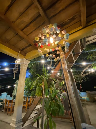 This image depicts an outdoor dining area at night, likely in Karimunjawa, Indonesia. The scene is illuminated by numerous string lights and a decorative light fixture hanging from a wooden beam. The fixture is a circular frame with colorful pom-poms and a glowing bulb at its center. A prominent white pillar with decorative molding is visible, suggesting the area is part of a building or a structured outdoor space. In the background, there are trees with lush green foliage, and wooden chairs and tables are arranged, indicating a restaurant or cafe setting. The overall atmosphere appears to be relaxed and inviting, with the warm glow of the lights creating a cozy ambiance. The time of day is clearly night, and the weather seems calm, suitable for outdoor dining. There are hints of people in the background, with one person's head and shoulders visible on the right side of the frame, appearing to be sitting or standing. The structure of the roof, made of wooden planks and beams, is also a noticeable detail.