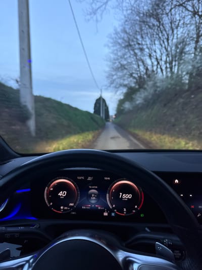 The image depicts a driver's perspective from within a vehicle, traveling on a narrow road in Tielrode, Belgium. The digital instrument cluster of the car, likely a Mercedes-Benz model, is prominent in the foreground. It displays a current speed of 40 km/h, an engine speed of 1500 r/min, an outside temperature of 12.0°C, and the time as 18:46. Trip information indicates "21 km" and "0:39" (minutes).
The road ahead is a single lane, appearing to be unpaved or made of worn asphalt, and is illuminated by the vehicle's headlights. To the left of the road, a grassy embankment rises, with a utility pole positioned close to the roadside. To the right, deciduous trees with bare branches line the road, interspersed with lighter-colored foliage, possibly early spring blossoms. The road extends into the distance, converging towards a cluster of dense trees. The sky is overcast, indicative of twilight conditions. No other vehicles or individuals are visible in the scene.