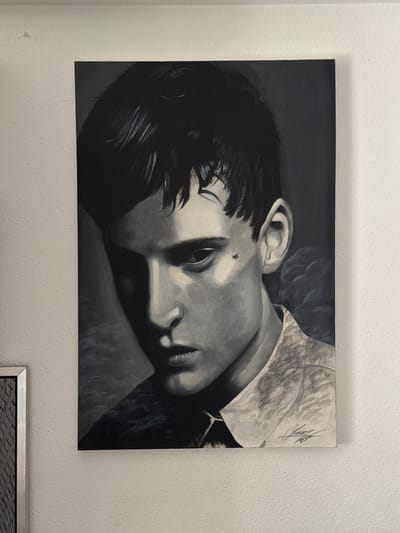 A black and white painting is displayed on a textured white wall. The painting is a close-up portrait of a young person lying down, with their head tilted and resting. The person has dark hair that falls across their forehead and cheek. Their eyes are partially closed, and their lips are slightly parted, giving a sense of repose or perhaps weariness. A small, dark mark, possibly a mole or a tiny insect, is visible on the person's cheekbone. The lighting in the painting creates strong contrasts, with dramatic shadows defining the facial features and the form of the head and neck. The person appears to be wearing a collared shirt, indicated by the fabric on the right side of the frame. The background of the painting is dark and indistinct, focusing all attention on the subject. On the upper right side of the painting, a handwritten signature in dark ink is visible. Below the painting, a small portion of a framed object with a metallic border and a patterned insert is visible. The context of Vigo, Spain, does not provide any specific visual cues within the image itself.