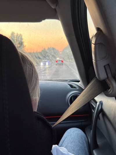 The image shows the interior of a car, viewed from behind the passenger seat. A person with blonde hair and wearing a black top and grey sweatpants is seated in the passenger seat, facing forward. A seatbelt is fastened across their chest. The car is on a road, with other vehicles visible in the distance. The sky is a warm, orange hue, suggesting it is either sunrise or sunset. The overall scene conveys a sense of travel and the ordinary.