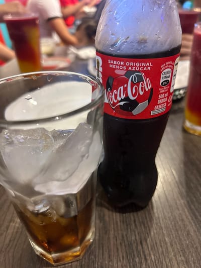 The image displays a close-up view of a Coca-Cola bottle and a glass with ice, positioned on what appears to be a wooden table. The image is rotated 90 degrees clockwise.

In the foreground, a dark-colored Coca-Cola plastic bottle lies horizontally. The left end of the bottle, containing what looks like ice or heavy condensation, is covered in a white, frosty layer, indicating it is very cold. The bottle's red label is prominently visible. The label features the iconic white "Coca-Cola" script logo, along with text in Spanish. Above the logo, it reads "SABOR ORIGINAL" and "MENOS AZÚCAR" (Original Flavor, Less Sugar). To the right of the main logo, a stylized soccer ball graphic is incorporated into the design, suggesting a special edition, possibly related to a sporting event. Further details on the label include nutritional information: "CALORIAS 150 kcal" (150 calories), "500 ml" (500 milliliters), "GASEOSA" (carbonated drink), and "BEBIDA" (beverage). There's also text stating "Si un vaso es de 200ml, en base a 2000 kcal" (If a glass is 200ml, based on 2000 kcal).

Next to the bottle, a clear glass stands upright, partially filled with large, irregularly shaped ice cubes and a dark brown liquid, presumably Coca-Cola. The glass reflects light, and its surface appears somewhat faceted. The dark liquid is visible beneath the ice and around the bottom of the glass.

The surface beneath the bottle and glass is a light brown wooden table, showing a distinct grain pattern. In the blurred background, other elements are visible, including what looks like other drinks in glasses or bottles with red and orange liquids, and indistinct shapes that could be people or other objects in a social setting, such as a restaurant or gathering.

The scene suggests a casual setting where a cold beverage is being served or enjoyed. The condensation on the bottle and the ice in the glass emphasize the chill of the drink.