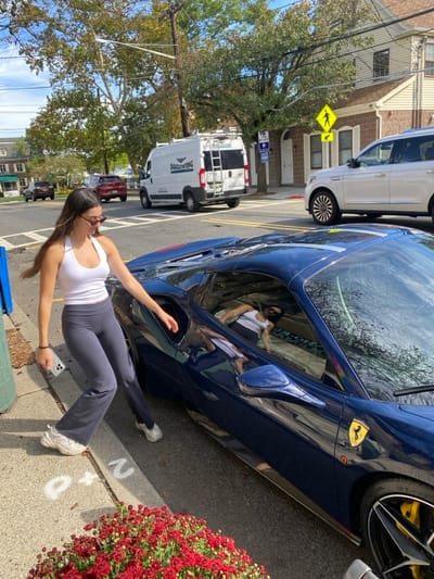 The image depicts a daytime street scene in Ridgewood, New Jersey, United States. In the foreground, a female with long dark hair, wearing a white halter top, dark gray flare pants, white sneakers, and sunglasses, stands on a concrete sidewalk. She holds a light-colored smartphone in her left hand and extends her right arm towards the open driver's side window of a dark blue Ferrari sports car, which is parked parallel to the curb. Inside the Ferrari, a reflection shows an individual, likely male, in a white tank top and dark sunglasses, reclining in the driver's seat. The Ferrari features yellow brake calipers and a yellow Ferrari emblem.

The immediate foreground includes a dark green utility pole and a cluster of red flowers in a planter. White chalk markings "P" and "2" are visible on the sidewalk. In the background, a white Ram ProMaster commercial van with "PACKWING" branding is present on the street. A white Lincoln Navigator SUV is parked or moving behind the Ferrari, and other vehicles, including a red SUV and a dark sedan, are visible further down the street. Residential-style buildings, utility poles with wires, blue street signs, and a yellow pedestrian crossing sign are present along the street, which features white crosswalk markings. The scene is brightly lit by natural sunlight, indicating clear weather conditions.