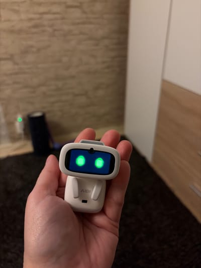 A person's hand holds a small white robot with a screen displaying two green eyes. The robot has the name "AIBI" printed on its lower body. In the background, a wall with a textured pattern is visible, along with a blurred white and wooden cabinet or door. A dark cylindrical object and a small green light are also present in the background. The scene appears to be indoors, likely in a domestic setting. The city of Härryda, Sweden, is not depicted or suggested in the image.