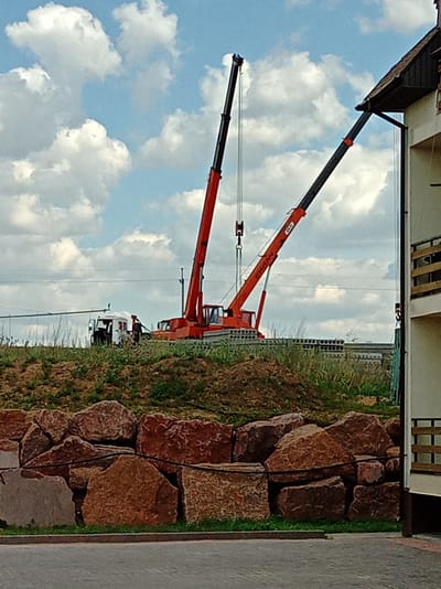 In Voronivka, Ukraine, a construction scene unfolds beneath a sky dotted with white clouds. Two large orange and black cranes dominate the frame, their booms extended upwards, suggesting ongoing work. One crane, positioned to the left, is the primary focus, with its cab visible. To its right, a second, larger crane is also at work. In the middle ground, a white truck with a partially visible cabin sits on a grassy incline. The area is marked by a low wall constructed of large, rough-cut reddish-brown rocks. Behind the wall, a series of gray concrete slabs with circular perforations are laid out, suggesting building materials. The background consists of a faint power line and a distant, sparsely vegetated horizon. On the far right, a portion of a building with a dark roof and white siding is visible, featuring a dark-colored balcony with horizontal railings.