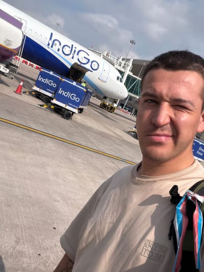 A man is taking a selfie at an airport in Vikram Nagar, India. He is wearing a beige t-shirt and has short dark hair. In the background, an Indigo Airlines airplane is visible, along with luggage carts and airport infrastructure. The sky is partly cloudy.
