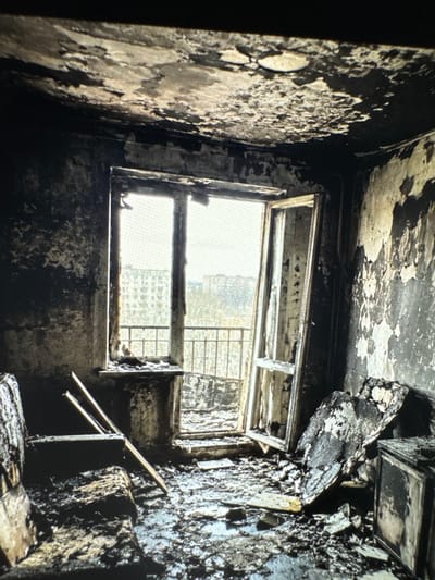 The image depicts the interior of a room in a residential building in Neubrandenburg, Germany, that has been severely damaged by fire. The ceiling shows significant charring and peeling plaster, indicative of intense heat exposure. The walls are blackened and stained, with large areas where the plaster has deteriorated. Debris, including charred wood and what appears to be melted or burnt material, covers the floor. In the foreground, the remnants of furniture, possibly a sofa and a cabinet, are visible, heavily damaged and coated in ash and soot. A large, burnt piece of material, possibly part of a wall or ceiling, lies on the floor near the center of the room. A window, partially open, looks out onto an urban landscape with apartment buildings and trees, suggesting the room is in a multi-story structure. The window frame itself appears charred and damaged. The overall scene is one of complete devastation and destruction caused by a fire.