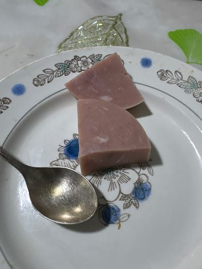 The image displays a close-up, overhead view of a white ceramic plate with a floral pattern, set against a light-colored, textured surface. On the plate, two triangular pieces of processed meat product, likely bologna or a similar cold cut, are positioned in the upper right quadrant. To the left of the meat, a silver spoon rests on the plate, its surface reflecting light. The floral pattern on the plate features stylized flowers with brown outlines and accents of light blue. The background shows hints of other decorative elements, including a green leaf-like shape and a shimmering, leaf-shaped object. The overall setting appears to be indoors, with soft lighting. The location is identified as the city of Siniy Gay, Russia. There are no people visible in the image, and no actions are being performed.