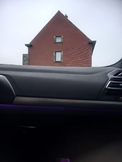 The scene displays the gable end of a red-brick house in Zwijndrecht, Belgium, viewed from inside a vehicle. The house exhibits a steeply pitched red-tiled roof with a central chimney. Two rectangular windows, each featuring white horizontal blinds, dark frames, and rough-hewn stone surrounds, are visible on the brick facade; the upper window is positioned higher than the lower. An architectural projection with dark tiling and a dark overhang is present on the left side of the house, while a smaller dark overhang is visible beneath the roofline on the right. The exterior environment consists of an overcast, grey sky. The foreground comprises the dark grey dashboard of the observing vehicle, bisected horizontally by a light-colored trim strip. A thin purple ambient light strip runs beneath the trim. A partial view of a silver-colored air vent is visible on the far right of the dashboard. No individuals or dynamic actions are present.