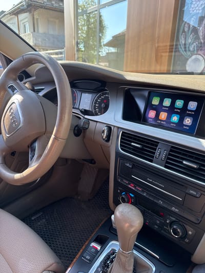 The image depicts the interior of an Audi car. The steering wheel, dashboard, gear stick, and infotainment screen are visible. The screen displays icons for AutoLink, Setup, AirPlay, Home, Android, USB Media, iOS, and DVR. The dashboard includes controls for climate control and multimedia. Outside, the view through the windshield reveals a building, suggesting an urban or suburban environment. Although the city is given as Pirdop, Bulgaria, there are no specific details in the image confirming this location.