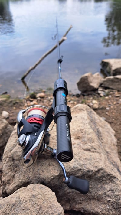 This media file depicts a fishing rod and reel placed on rocks next to a body of water. The fishing rod, with a black carbon fiber handle, has the word "FAVORITE" and "nano" printed on it. A fishing reel with a red and silver spool is attached to the rod. The scene is set outdoors, with the water reflecting the sky, suggesting it's daytime. The presence of rocks and what appears to be a bank indicates a natural environment, likely a lake or river. The image is shot from a close-up perspective, focusing on the fishing equipment. There are no people visible in the frame. The time of day appears to be either morning or afternoon, with the lighting not harsh enough for midday. The weather seems calm, as the water's surface is relatively smooth. The location context provided is Krosno Odrzańskie, Poland.