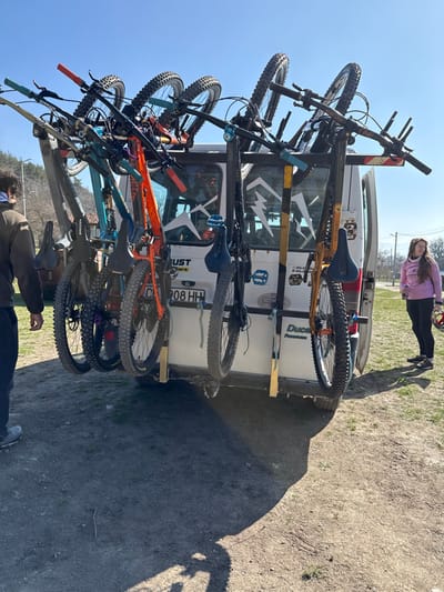 In the city of Tsarkvishte, Bulgaria, a white van equipped with a multi-bicycle rack on its rear is parked on an unpaved dirt surface. Six mountain bicycles of varying colors (orange, turquoise, dark grey, and yellow-orange) are mounted vertically on the rack. The van displays a Bulgarian license plate "C 908 HH" and partial "DUCA" and "Passa" branding on its lower right rear. Stickers, including a large white mountain silhouette, are visible on the rear windows.
To the left, a male individual, partially visible from the back, wears a dark long-sleeved top, dark trousers, and light grey shoes, facing towards the van. To the right, a female individual stands, dressed in a pink long-sleeved hooded top with white accents and black leggings, holding a red and white bicycle helmet in her left hand. She looks towards the van.
The scene is under a clear blue sky, with bright sunlight casting prominent shadows of the van and bicycles onto the ground. In the background, deciduous trees are visible to the left, and a utility pole with wires is present to the far right. The ground consists of dirt with sparse patches of green grass.