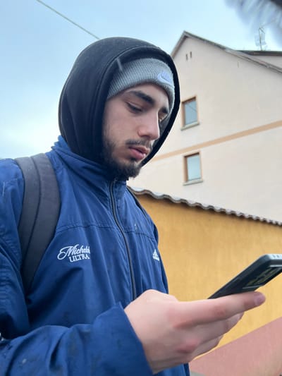 The image depicts a young adult male outdoors in what is identified as Meistratzheim, France, under overcast weather conditions with light precipitation. The male, positioned in the foreground and framed from the chest up, is seen in profile, looking down at a smartphone held in his right hand. He wears a grey knitted beanie with a white Nike logo, covered by the hood of a black sweatshirt. Over this, he wears a navy blue athletic jacket featuring a white "Michelob ULTRA" logo on the left chest and an Adidas logo on the right chest. A black backpack strap is visible over his right shoulder. His right thumb is positioned over the phone screen, suggesting active interaction. The background features two buildings: an upper structure with a light peach-colored facade and two rectangular windows, topped by a brown tiled roof, and a lower structure with a yellow facade and a visible brown tiled roof. Small white specks, consistent with light snow or sleet, are visible on his clothing and in the air.