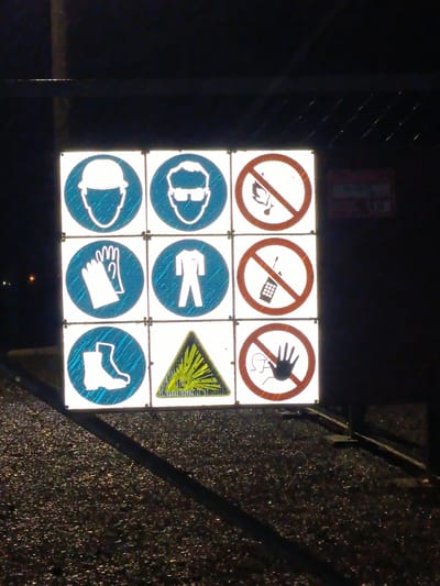 A grid of nine reflective safety signs is mounted on a fence. The signs are arranged in three rows and three columns, with each sign depicting a different safety symbol or prohibition. The top row shows a hard hat, safety glasses, and a no smoking sign. The middle row displays a pair of gloves, protective clothing, and a no mobile phone sign. The bottom row features safety boots, a warning sign for explosive materials, and a no touch sign. The ground beneath the signs is dark and appears to be gravel or asphalt, with reflections from the lights visible on its surface. The background is dark, suggesting nighttime or a poorly lit area. A metal fence runs horizontally across the top of the image, with faint lights visible in the distance. The overall scene is a clear depiction of safety regulations being communicated through standardized signage.