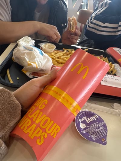 The image depicts a meal at McDonald's, likely located in Portadown, United Kingdom. A "Big Flavour Wraps" container is held in a person's hand, the McDonald's logo visible. The table has a tray with fries, a wrapped burger, and a container of garlic mayonnaise. Another person, partially visible, holds a bite-sized burger. A second tray is on the table, marked "ER 42, Crispy Wrap, Time 13:59."