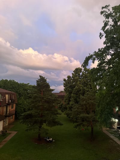 The image captures a dusky sky over an urban residential area. In the foreground, two large pine trees stand on a grassy lawn, with a white all-terrain vehicle parked near the base of the right tree. Adjacent to the lawn, a brick apartment building with brown roofs and balconies is visible on the left, and another similar building is partially seen on the right. Dense green foliage surrounds the buildings and the courtyard. The sky is filled with dramatic clouds, illuminated by the fading light of dusk, with hints of pink and purple hues. The scene appears to be a quiet evening in a suburban setting, likely in Glendale Heights, USA.