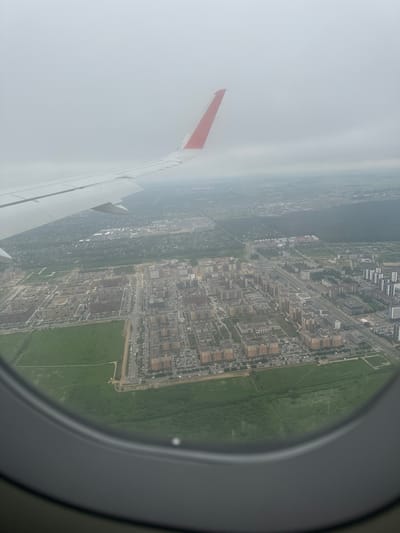 This is an aerial photograph taken from an airplane window, looking down on the city of Slavyanka, Russia. The image is characterized by a hazy, overcast sky, which contributes to a muted color palette. The aircraft's wing, painted white with a red tip, occupies the upper left portion of the frame, extending diagonally across the sky.
Below the wing, a dense urban landscape unfolds. Numerous apartment buildings, characterized by their reddish-brown facades and rectangular shapes, are visible. These buildings are clustered together, forming distinct neighborhoods. Streets and roads weave through the cityscape, with cars parked along them, indicating a populated area.
Adjacent to the built-up areas are large expanses of green, likely parks, fields, or undeveloped land. These green spaces provide a contrast to the man-made structures. In the distance, more buildings and greenery can be seen, fading into the hazy atmosphere. The overall impression is of a modern, developing city under a gray, cloudy sky. The view is framed by the circular airplane window, which occupies the bottom portion of the image.