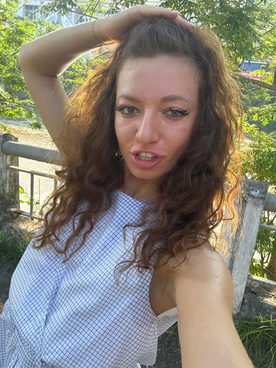 A woman with long, wavy brown hair is taking a selfie outdoors. She is wearing a light blue and white checkered shirt and is touching her hair with her left hand. The background includes greenery, buildings, and a concrete structure. Based on the location listed, the scene appears to be in a residential or mixed-use area of Lao Cai Ward, Vietnam.