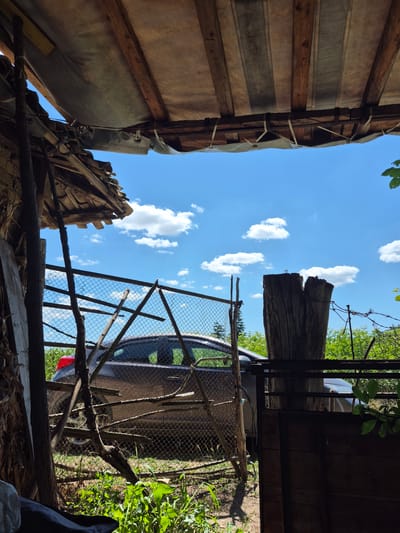 The image depicts an outdoor scene, likely a yard or rural area, under a bright blue sky with scattered white clouds. In the foreground, a makeshift shelter is visible, constructed from wooden beams and a tarp. To the left, a pile of wooden planks leans against a vertical support. A chain-link fence runs across the midground, partially obscuring a brown car. The car appears to be parked. Beyond the fence, lush green vegetation is abundant, suggesting a natural or overgrown environment. A thick, weathered wooden post stands prominently to the right of the fence, with nails visible on its surface. The overall impression is one of a rustic and somewhat unkempt setting. The specific location is identified as Sadina, Bulgaria.