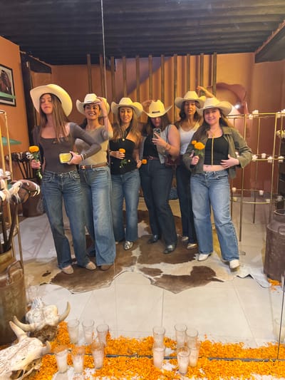 Six adult females, all wearing cowboy hats and jeans, are depicted indoors in Guanajuato City, Mexico, posing for a photograph. The setting features a rustic, western-themed decor with elements suggestive of a cultural celebration.

From left to right, the women are dressed in varying tops: a dark brown long-sleeve top, a grey long-sleeve top, a black halter-neck top, a black top with a grey vest, a white tank top, and a black top with an olive-green short jacket. All wear blue jeans of different washes and styles, with footwear ranging from flats to white cowboy boots. Three of the women hold single orange marigold flowers. One woman on the left holds a small rectangular object, possibly a phone. Another woman near the center right holds a white smartphone to her face.

The room features orange-toned walls, a dark ceiling with visible beams, and a prominent wooden slatted wall feature in the background. The floor is light-colored tile, partially covered by cowhide rugs. Decorative elements include: a multi-tiered metal stand holding multiple lit candles in glass holders on the right; two large, rusted metal milk cans (one on the far left, one on the far right); and a framed picture on the left wall. In the immediate foreground, separated by a clear barrier, two animal skulls with horns are arranged on the floor amidst a dense scattering of orange marigold petals and several empty clear drinking glasses. A large mirror or reflective surface is present, reflecting portions of the scene.