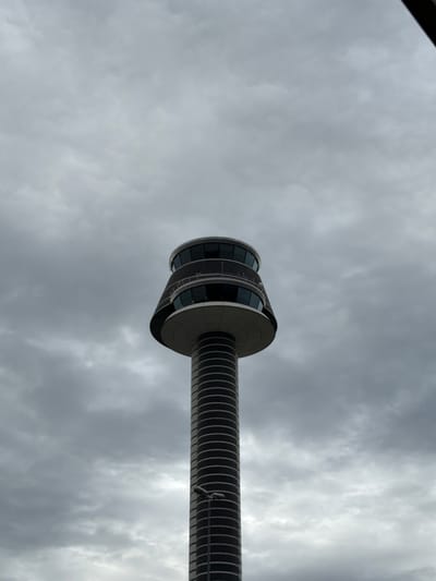 The image shows the Stockholm-Arlanda Airport Air Traffic Control Tower against a cloudy sky. The tower is a tall, cylindrical structure with a distinct observation deck at the top, featuring multiple large windows. The deck is circular and wider than the main body of the tower, which is ribbed or segmented. A street light is visible on the side of the tower in the lower portion of the frame. The overall atmosphere is overcast and grey. There are no people visible in the image.