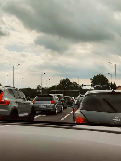 The image depicts a traffic jam on a cloudy day in Sławków, Poland. Multiple cars are stopped on a multi-lane road. In the foreground, the rear of a silver Volvo SUV is visible, followed by a silver Opel hatchback with license plate "WPN 00483." Several other vehicles are in the background, also stationary. Traffic lights are present, with some indicating red and others green. Streetlights line the side of the road, and trees are visible in the distance. A road sign indicating a U-turn prohibition is also visible. The sky is filled with heavy, gray clouds.