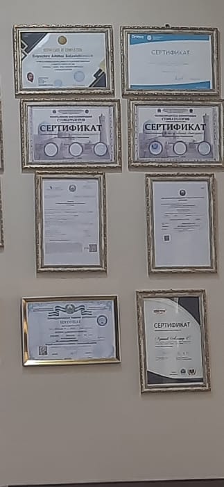 The scene depicts the interior of a professional establishment, likely a dental clinic, located in Kogon City, Uzbekistan. Nine framed documents, predominantly certificates, are displayed vertically on a light-colored, plain wall. Each document is encased in an ornate, gold-colored rectangular frame with decorative borders.
Several certificates feature the Cyrillic word "СЕРТИФИКАТ" (Certificate). Two prominent certificates in the second row from the top include an illustration resembling a building or structure with four circular elements, and the Cyrillic word "СТОМАТОЛОГИЯ" (Stomatology/Dentistry), confirming a dental context. The top-left certificate shows a small photographic portrait of an individual and partially visible English text "CERTIFICATE OF COMPLETION" alongside the name "Ergashev Akmal Eshkabulu...". Other documents display various logos, seals, and QR codes. No individuals are present, and no actions or interactions are depicted. The lighting is uniform, illuminating the displayed documents against the neutral wall surface.