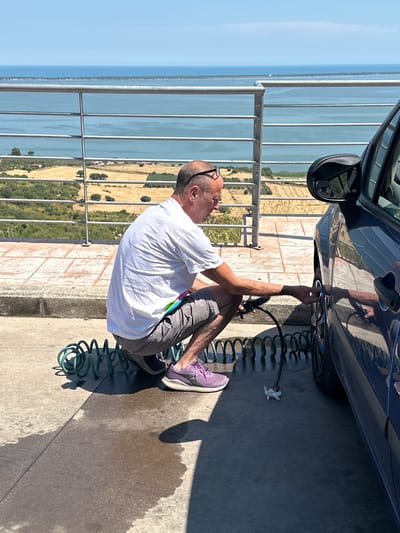A middle-aged Caucasian man with short, graying hair and sunglasses perched on his head is crouched down next to a dark blue car, washing its wheel with a black hose that is coiled on the ground. The man is wearing a white t-shirt and gray shorts. The car is positioned on a paved area, and a coiled green hose is visible on the ground. In the background, there is a body of water, possibly the Gargano coast of Italy. The man appears to be in a residential area or a scenic overlook, as evidenced by the railing and the expansive view of the water and landscape. The sun is shining brightly, casting shadows on the ground.
