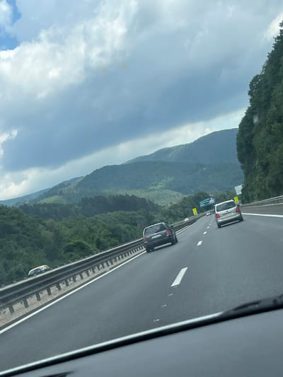 The image depicts a view from a vehicle traveling on a multi-lane highway with a mountainous, forested landscape on the left and a dense forest on the right. The sky is overcast with dramatic clouds. Several cars are visible on the road, including a dark sedan in the left lane and a silver minivan in the right lane, both moving away from the viewer. A guardrail separates the highway from the embankment on the left. In the distance, signs for a rest stop or advertisement can be seen. The scene is consistent with a highway in a mountainous region of Churek, Bulgaria, characterized by its natural beauty and infrastructure.