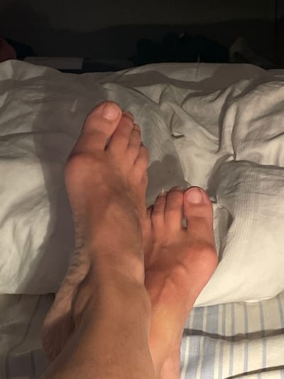 A close-up, low-angle shot shows two bare feet resting on a wrinkled white duvet. The feet are illuminated from the front, casting shadows behind them. The left foot is positioned slightly above and in front of the right foot. The toes are clearly visible, with short, clean nails. The skin tone is warm and appears to have some redness, possibly from tanning or irritation. The background is dark and out of focus, with hints of bedding and possibly furniture. The scene appears to be indoors, in a bedroom. The location in Oldenburg in Holstein, Germany is not discernible from the visual information provided in the image.