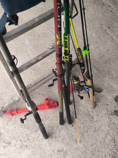The image displays a collection of fishing rods and associated gear, leaning against a weathered wooden ladder or similar support structure. The items are situated outdoors on a light grey, textured concrete or asphalt surface.

Several fishing rods are visible, showcasing various designs and colors. A prominent rod, dark red or maroon, features bright yellow text reading "SION 500" and "S.00.01.15" on its lower section. Another rod is green with a yellow tip and a bright green securing clip. A third rod has a distinctive camouflage-patterned handle. Most rods are equipped with fishing reels; at least two spinning reels are discernible, one dark grey/black with a crank handle, and another with a silver spool clearly showing "5000" and fishing line. A black rod near a bright green clip bears the text "SPINNING." One rod has a cork handle, while another has a bright orange sensitive tip.

On the ground, behind some of the rods, lies a vivid red, plastic hand-operated ice auger, strongly suggesting an involvement with ice fishing. The wooden support structure appears aged, with horizontal rungs.

The scene is brightly lit, indicative of daytime under clear weather conditions. The arrangement of the gear implies storage, organization, or preparation for a fishing excursion. No individuals are present in the frame. The location context provided is Dolistovo, Bulgaria.