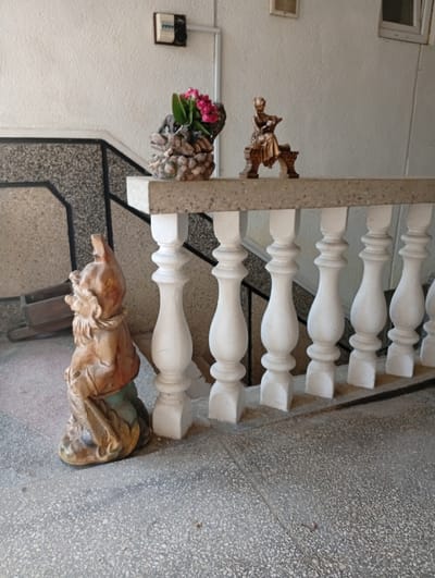 The image shows a scene likely located in Veliatyno, Ukraine, on an outdoor stairway with a concrete railing. To the left, a garden gnome statue is seated on the ground, facing right. The gnome is depicted with a red hat and green trousers. Adjacent to the gnome, on a small concrete pedestal, is a decorative pot filled with pink flowers and greenery. Further along the railing, a small bronze-colored statue of a woman seated on a bench can be seen. The stairway itself is made of concrete, with a speckled gray surface. The wall behind the railing is off-white. A wooden cart is visible in the background to the left, and electrical fixtures are mounted on the wall.