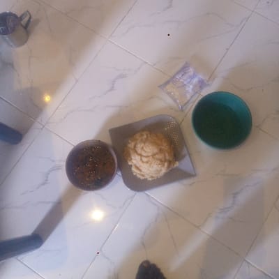 The image displays an indoor scene on a light-colored, white and grey marble-patterned tiled floor in Awo Ekiti, Nigeria. A meal is set out, featuring a brown, rounded bowl on the left containing a dark, reddish-brown stew or soup with visible green and red components. Adjacent to it on the right, a light grey, rectangular plate with rounded corners holds a large, textured mound of a light beige, solid food, consistent with a 'swallow' dish. An empty, dark teal-colored plastic bowl is positioned to the right of the plate. Above this empty bowl, a clear plastic sachet, likely containing water, rests on the floor. In the upper left quadrant, a silver-colored metal mug with a handle is visible. A dark, partially obscured object, possibly a furniture leg, is present on the far left edge. A bare, dark-skinned human foot is partially visible in the bottom right corner of the frame, indicating the presence of an individual. Multiple bright reflections from an unseen light source are visible on the tiled surface.