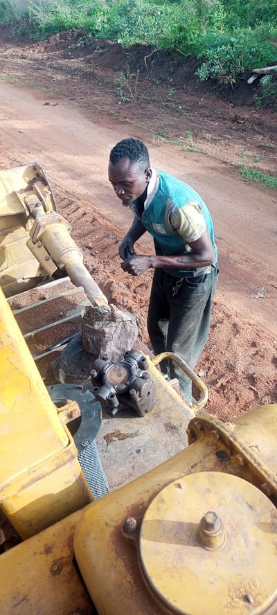 This image captures a focused man performing maintenance or repair work on heavy machinery in an outdoor setting in Otunade, Nigeria.

The central subject is an adult man with short, dark, textured hair and a beard. He is wearing a short-sleeved t-shirt, predominantly teal or light blue with light yellow or cream sections on the shoulders and sleeves, and dark trousers. A set of keys hangs from a loop on his pants. He is leaning over, intently examining or working with his hands on a component of the machinery.

The machinery itself is large and yellow, likely a bulldozer or excavator, evidenced by the visible tracks in the background and a prominent hydraulic cylinder extending from its frame. Resting on a flat surface of the machinery are mechanical parts, including a universal joint (a cross-shaped coupling) and a round metal plate, indicating ongoing mechanical work. On a circular yellow cap towards the bottom right of the frame, the text "BJ926" is faintly visible.

The setting is an unpaved area with reddish-brown earth, which appears to be a typical rural or construction site. In the background, a natural bank of green foliage and disturbed soil completes the scene. The lighting suggests it is daytime, either morning or afternoon, with clear weather conditions. The man's expression is one of concentration and diligence as he works on the equipment.