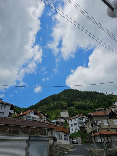 The image depicts a view in Erma Reka, Bulgaria, looking towards a hillside dotted with houses and dense green trees. The sky is visible, with patches of blue interspersed with white and gray clouds. Power lines are present in the foreground, stretching across the sky. The houses on the hillside vary in size and design, some with red-tiled roofs and others with more modern architecture. The scene captures a typical residential area nestled in a natural setting.