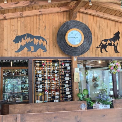 The image depicts an interior scene at a location identified as "Atoluka, Bulgaria." The environment is characterized by exposed wooden beams and vertical wooden paneling on the walls and ceiling. On the wall, there are two silhouette art pieces: one of a bear with trees inside its silhouette on the left, and another of a wolf howling with trees inside its silhouette on the right. A large, circular wicker-framed clock with a white face and black Roman numerals is centrally located between the two silhouettes.

Below the wall art, a section of the establishment features a large glass-fronted cabinet filled with various bottles of liquor and spirits. Adjacent to this cabinet is a wall-mounted wine rack holding numerous bottles of wine. Some of the wine bottles have decorative elements, such as small mushroom-shaped ornaments.

In the foreground, a wooden counter or bar area is visible. The overall impression is that of a rustic-themed establishment, possibly a bar, restaurant, or winery, with decorative elements that evoke a natural or wilderness theme. No people are visible in the image.