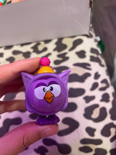 An extreme close-up shot shows a person's thumb and index finger holding a small, purple owl figurine. The owl has large white eyes with black pupils, an orange beak, and a yellow hat topped with a pink, textured ball. The background is a blurry, black and white spotted pattern, possibly a fabric. The image is taken from a low angle. There is no indication of Borisova Griva Massiv, Russia.