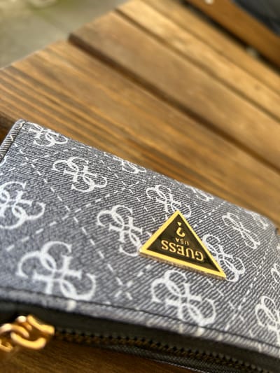 The image captures a close-up, angled shot of a patterned wallet or small purse resting on a wooden surface. The wallet features a greyish-blue fabric with a repeated white monogram pattern, appearing to be interlocking letters, possibly "G" or "C" shapes, interspersed with dashed lines that suggest a denim-like texture. A prominent golden triangular emblem with a black background is affixed to the wallet. Clearly visible on this emblem is the word "GUESS" in gold letters, positioned vertically along one side of the triangle, with "USA" and a question mark symbol also in gold. A dark zipper with a small golden pull tab is visible along the right edge of the wallet.
The background consists of blurred wooden planks, suggesting an outdoor setting or a piece of wooden furniture like a table or bench. The wood has a warm, light brown tone with visible grain. The lighting indicates it is daytime, with natural light illuminating the scene. No people are visible. The object is stationary, implying no specific activity other than being placed on the surface. While the exact location is not discernable from the image alone, the wooden setting could be consistent with outdoor areas at Gara Elin Pelin, Bulgaria, such as a bench at a train station or a cafe table.