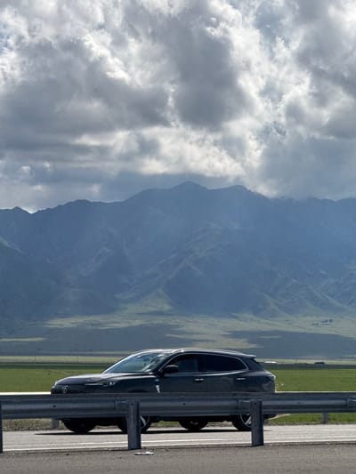 The image displays a serene mountainous landscape observed from a roadside vantage point. In the foreground, a dark-colored SUV or crossover vehicle is parked on an asphalt road shoulder, positioned next to a metallic guardrail. A faint circular logo on the vehicle's front suggests it might be a Nissan. The vehicle faces towards the right, with its front wheels visible below the guardrail.
Beyond the road and guardrail, the midground features expansive, verdant green fields that gradually ascend towards the base of a substantial mountain range. These fields exhibit subtle variations in texture and color, hinting at natural contours. In the background, majestic mountains dominate the horizon, appearing in muted blue-grey tones, indicating significant distance and atmospheric haze. Their imposing forms extend upwards, with peaks partially obscured by the soft lighting.
The sky is filled with a mix of white and grey clouds, characteristic of a partly cloudy daytime. Some areas of the clouds are brighter, suggesting sunlight is breaking through and illuminating the scene. The overall ambiance is one of natural beauty and tranquility. No people are visible. The location context provided places this scene in Otar Rgaytö, Kazakhstan.