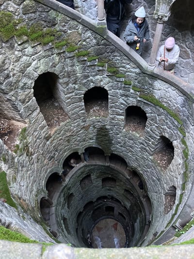 This image depicts the vertical descent into the Initiation Well (Poço Iniciático) at Quinta da Regaleira, located in Sintra, Portugal (specifically within Santa Maria e São Miguel, São Martinho e São Pedro de Penaferrim). The perspective is an overhead view looking directly down into the approximately 27-meter deep dry well, which is constructed from irregular, grey, rough-hewn stone blocks, heavily textured and showing patches of green moss adhering to surfaces, particularly on the upper levels and within arched recesses.
A continuous stone spiral staircase with stone railings and decorative balusters winds clockwise down the entire interior of the conical well structure. At various points along its descent, multiple semicircular arched openings lead into dark tunnels or chambers within the surrounding rock and earth. The ground visible within these openings consists of loose soil and scattered dry leaves.
At the uppermost visible section, three individuals are present. One person, largely obscured on the left, wears dark clothing. To their right, a person with dark hair, wearing a grey beanie and a dark green jacket, holds a dark electronic device (likely a smartphone or camera) with both hands, looking downwards. A yellow lanyard is visible around their neck. Further right, another person with light hair, wearing a light pink beanie and a light-colored jacket, similarly holds a dark electronic device with both hands, also focused downwards.
Midway down the well, on the descending staircase, one individual in dark clothing with a reddish hat or hood is visible leaning forward, appearing to navigate the steps. At the very bottom of the well, a circular paved area is visible, appearing somewhat wet. A faint geometric pattern, possibly a cross or compass rose, is discernible in the center of the base. A solitary figure stands at the bottom of the well, their details indistinct due to distance and lighting. The overall lighting transitions from moderate brightness at the top to deep shadow at the bottom, illuminating the upper sections more clearly.