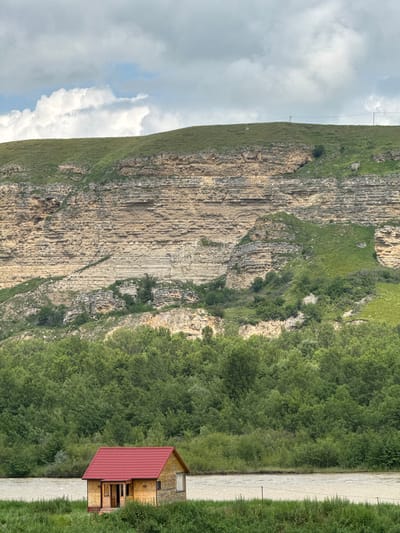 The image depicts a serene natural landscape with a small, wooden house featuring a red roof situated in the foreground. The house is located on the bank of a wide, murky river, bordered by lush green grass and dense trees. In the background, a massive, layered rock formation dominates the scene, its stratified layers indicative of geological history. The rock face is partially covered in green vegetation, and the sky above is overcast with dramatic clouds. The setting appears to be a rural or wilderness area, possibly in the vicinity of the village of Kubina, Russia, given the natural terrain and architecture. There are no people or animals visible in the image, and the environment suggests a tranquil, untouched environment.