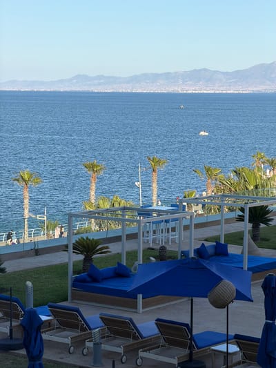 The image shows a coastal scene in Nador, Morocco, featuring a seaside resort or private residence. Visible are palm trees lining a balcony or terrace overlooking the Mediterranean Sea. Sun loungers with blue cushions and matching umbrellas are arranged on the terrace, indicating a relaxation area. The sea stretches towards the horizon, with mountains visible in the distance. Boats are scattered across the water, adding to the nautical scene. The setting suggests a sunny day with calm sea conditions.