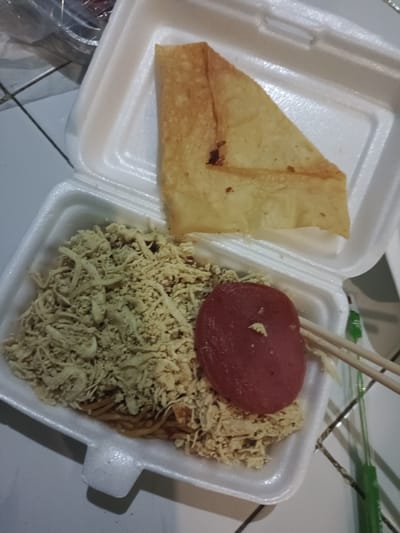 A white styrofoam food container is centrally positioned on a light-colored tiled floor with dark grout lines. The main compartment on the left contains a meal primarily consisting of noodles, largely covered by a mound of finely shredded, light brown material, consistent with shredded chicken or meat floss. A single, flat, circular slice of reddish-pink processed meat rests atop the shredded topping. To the right, a pair of light-colored wooden chopsticks is placed across the container. The smaller compartment, or lid, of the container holds a single, large, golden-brown, irregularly shaped fried pastry, possibly a fried wonton skin, which appears folded. Blurred transparent plastic bags or packaging are visible in the upper-left background, and a green plastic object is partially visible on the tiled floor to the far right. This scene is located in the city of Singopadu, Indonesia.
