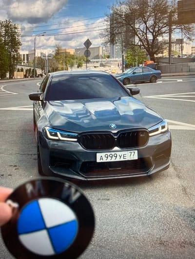 A dark gray BMW sedan with a license plate reading "A999YE 77 RUS" is parked on a city street. In the foreground, a person's hand holds a circular BMW emblem. The background shows buildings, trees, and a blue sky with clouds. The street is paved and appears to be in an urban environment. The image is taken from a slightly elevated perspective, looking down at the car.