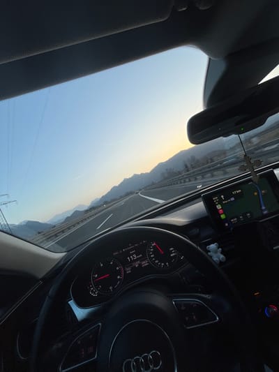 The scene depicts the interior of an Audi vehicle, viewed from the driver's perspective, traveling on a multi-lane highway near Ohlstadt, Germany. The time is 18:05 on March 7, 2024, with an outside temperature of -13.5°C and the vehicle's odometer reading 27228 kilometers.

The vehicle is moving at 113 km/h, as indicated by the speedometer on the instrument cluster. A central infotainment screen displays a navigation map showing "17 km" remaining to a destination and the current time of 18:05. A wooden crucifix hangs from the rearview mirror, and a small white teddy bear figurine is positioned on the passenger-side dashboard vent.

Outside the vehicle, a clear sky transitions from light blue overhead to a warm orange hue on the horizon, indicating late afternoon or early evening with the sun low. Mountains flank both sides of the highway, extending into the distance. Power lines and a large transmission tower are visible on the left side of the road. Other vehicles are discernible further down the highway.