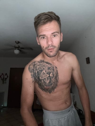 A shirtless man with a lion tattoo on his chest stands in a dimly lit room. He has light brown hair, brown eyes, and stubble. He is wearing grey sweatpants. In the background, a ceiling fan is visible. The man appears to be in a residential setting, possibly in Sesto Calende, Italy.
