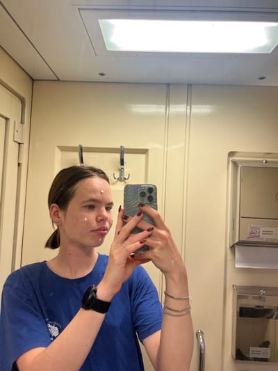 A young woman with brown hair pulled back in a ponytail and a blue t-shirt is reflected in a mirror. She is applying white cream to her face, with dots of cream visible on her forehead and cheeks. She is holding a smartphone in her right hand, taking a selfie. The background appears to be a train compartment, with cream-colored walls, a long fluorescent light fixture on the ceiling, and two hooks mounted on the wall. To the right of the woman, a metal dispenser with a label featuring a cigarette and a prohibition sign is visible. She is wearing a smartwatch on her left wrist and several thin bracelets on her right arm. The overall setting is likely a small bathroom or lavatory within a train. The image does not provide information about the city of Bessonovka, Russia.
