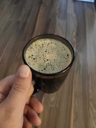A close-up shot shows a hand holding a dark glass mug filled with foamy coffee. The mug is dark brown, and the coffee has a light brown foam with small bubbles. The background is a wooden surface with a visible grain pattern, suggesting a table or floor. The lighting casts subtle shadows, indicating an indoor setting. The image does not contain any information about its location in Андрэеўшчынскі сельскі Савет, Belarus.