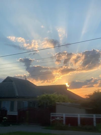 The image depicts a twilight sky with sunbeams radiating from behind clouds. Below the sky, a dark silhouette of houses and trees is visible. The scene appears to be a residential area, possibly on the outskirts of a city. There are no people or specific actions visible in the image. The overall mood is serene and captures the beauty of a sunset. This image is located in Svetlîi, Moldova.
